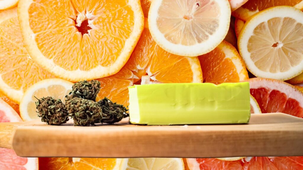 image of 4 buds of cannabis next to a block of butter on a wooden cutting board with a background of sliced citrus fruit for the post on cannabis infused citrus butter