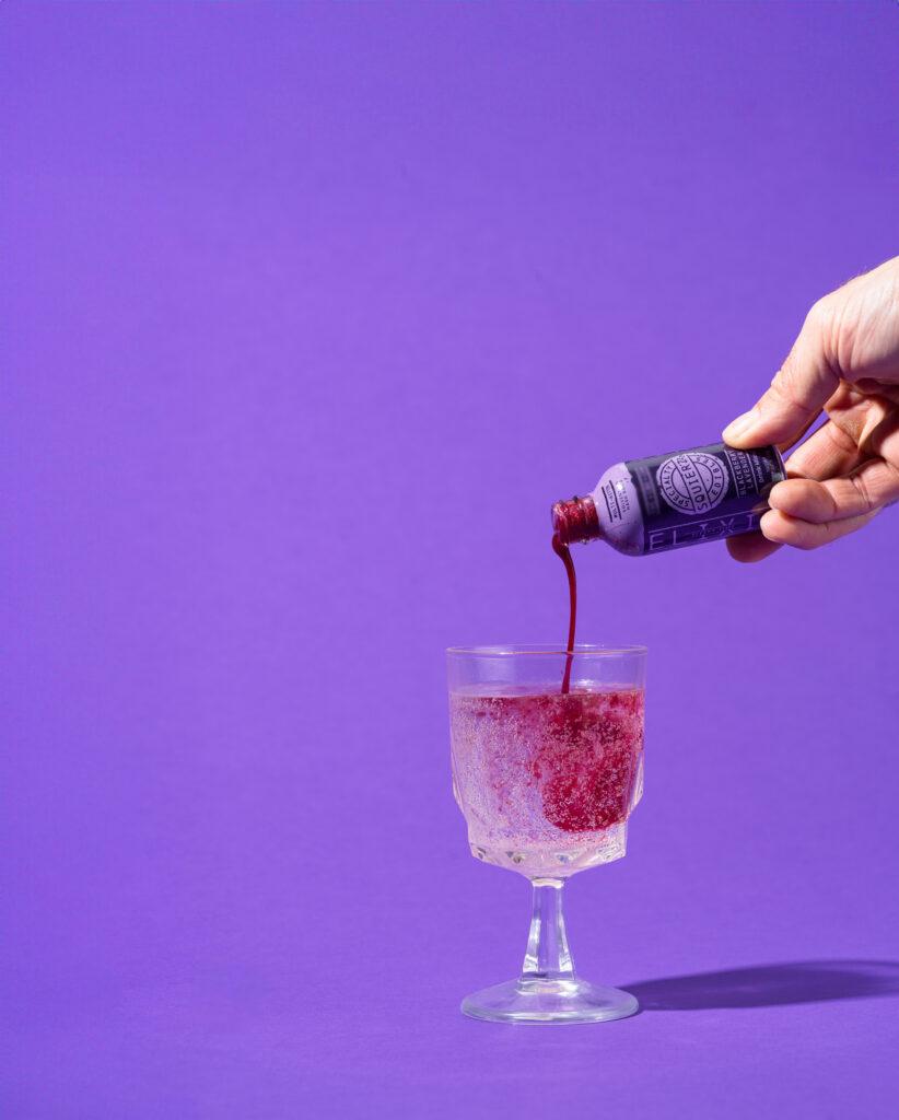 Squier's Specialty Elixirs purple bottle being poured into a wine glass full of a clear liquid