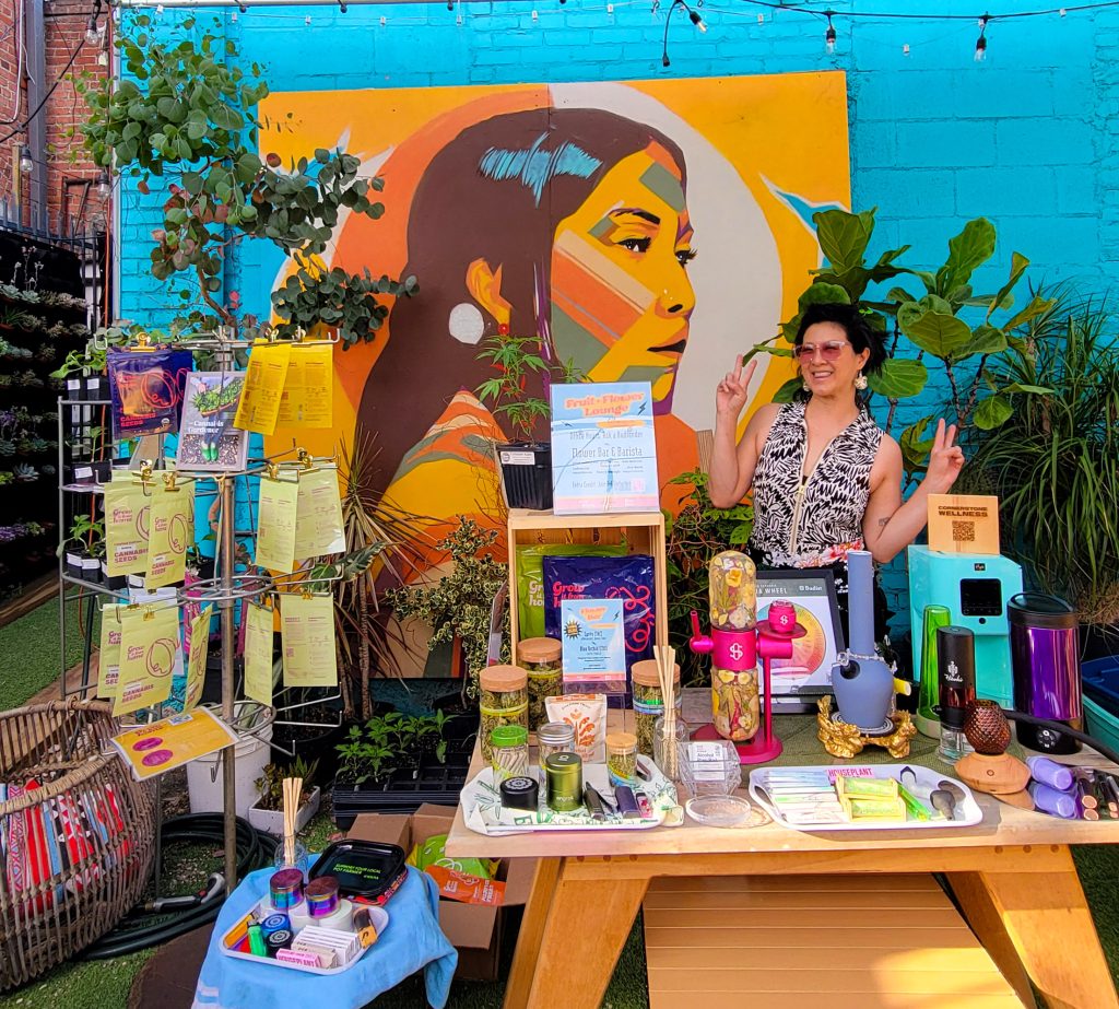 Christina Wong, founder of Fruit & Flower, giving peace signs behind a table at a outdoor cannabis.